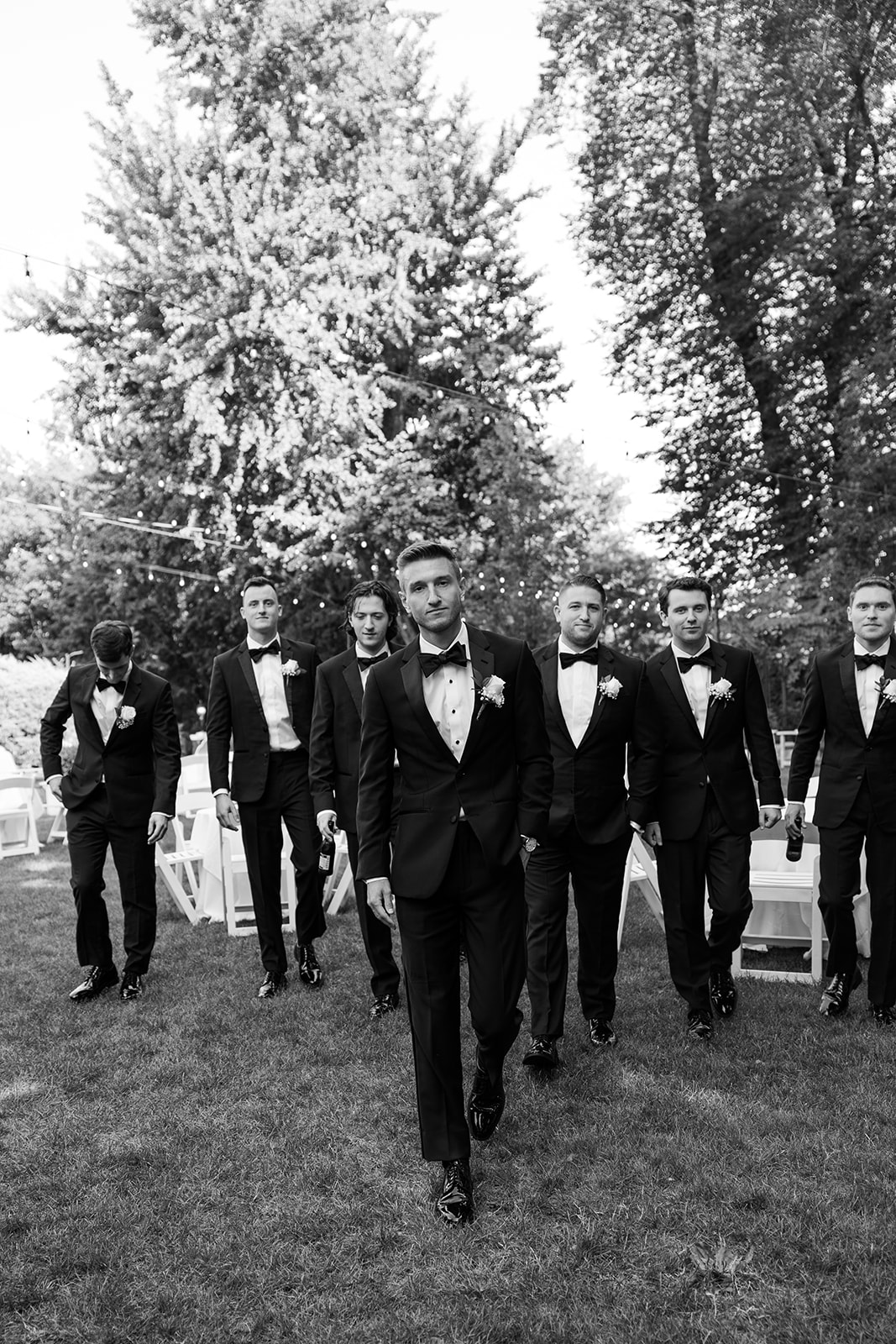 Black and white photo of the groom walking confidently forward on the lawn, followed by his groomsmen.